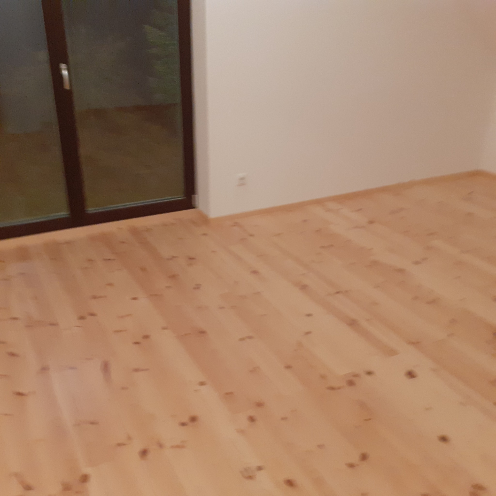 Jasny parkiet z widocznymi słojami drewna, ułożony w nowym wnętrzu z oknem balkonowym.