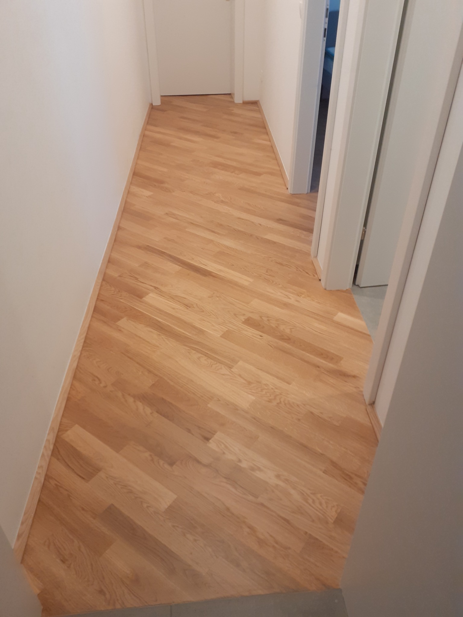 Ułożony parkiet w korytarzu, deski ułożone pod kątem, widoczne listwy przypodłogowe i białe drzwi.