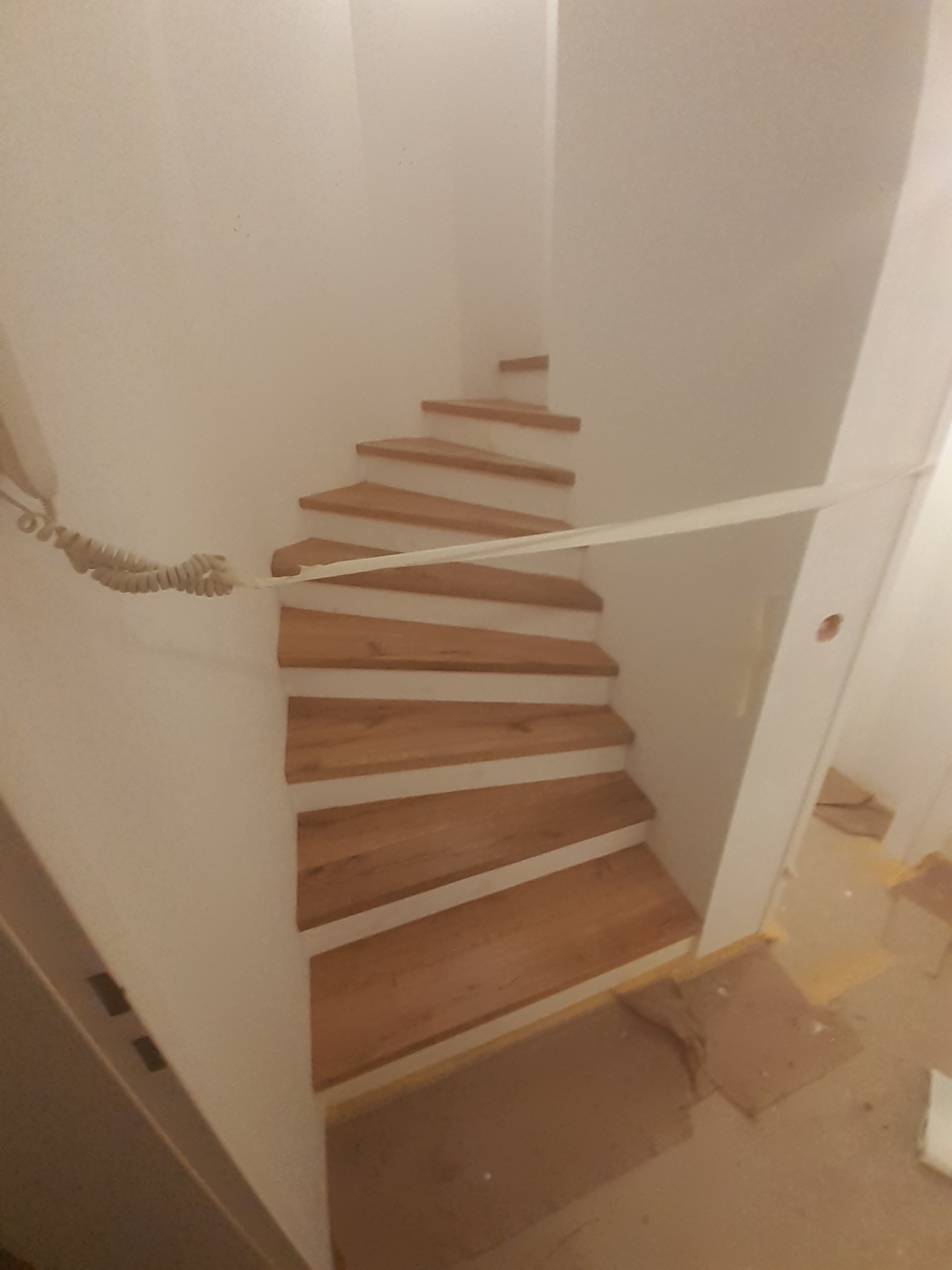 Wąskie, kręte schody z drewnianymi stopniami i białymi podstopnicami, zabezpieczone taśmą malarską, widoczne z góry w trakcie prac wykończeniowych.
