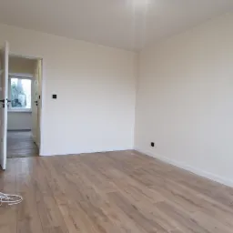 Jasne panele podłogowe ułożone w pustym pokoju z kremowymi ścianami, widoczne białe listwy przypodłogowe, drzwi wejściowe i oświetlenie sufitowe z trzema reflektorami.