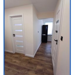 R.S.R. Radoslaw - Korytarz po remoncie z dwiema białymi drzwiami z czarnymi klamkami, panelami podłogowymi w odcieniach brązu oraz czarną lodówką w tle.