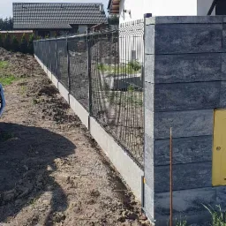 Nowoczesne ogrodzenie panelowe z betonowymi słupkami, świeżo osadzone w ziemi, z widocznym domem w tle.