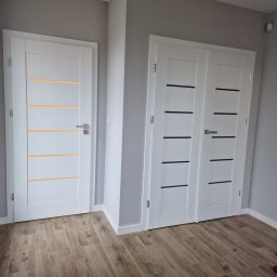 R.S.R. Radoslaw - Dwa rodzaje białych drzwi wewnętrznych z poziomymi frezami (żółte i czarne), zamontowane w szarej ścianie na tle podłogi z paneli imitujących drewno.