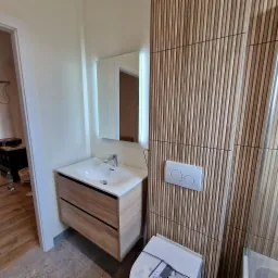 Nowoczesna łazienka po remoncie: drewniane lamele na ścianie, podświetlane lustro nad umywalką z szafką i widoczny fragment toalety. Stonowana kolorystyka.