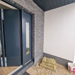 R.S.R. Radoslaw - Montaż drzwi zewnętrznych z kamienną okładziną elewacyjną. Widoczny etap prac, izolacja i niebieskie drzwi z szybą. Wykończenie w nowoczesnym stylu.