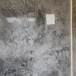 Ściana z efektem betonu architektonicznego w szarościach, oświetlona pionową listwą LED, z widocznymi dwoma białymi puszkami instalacyjnymi.