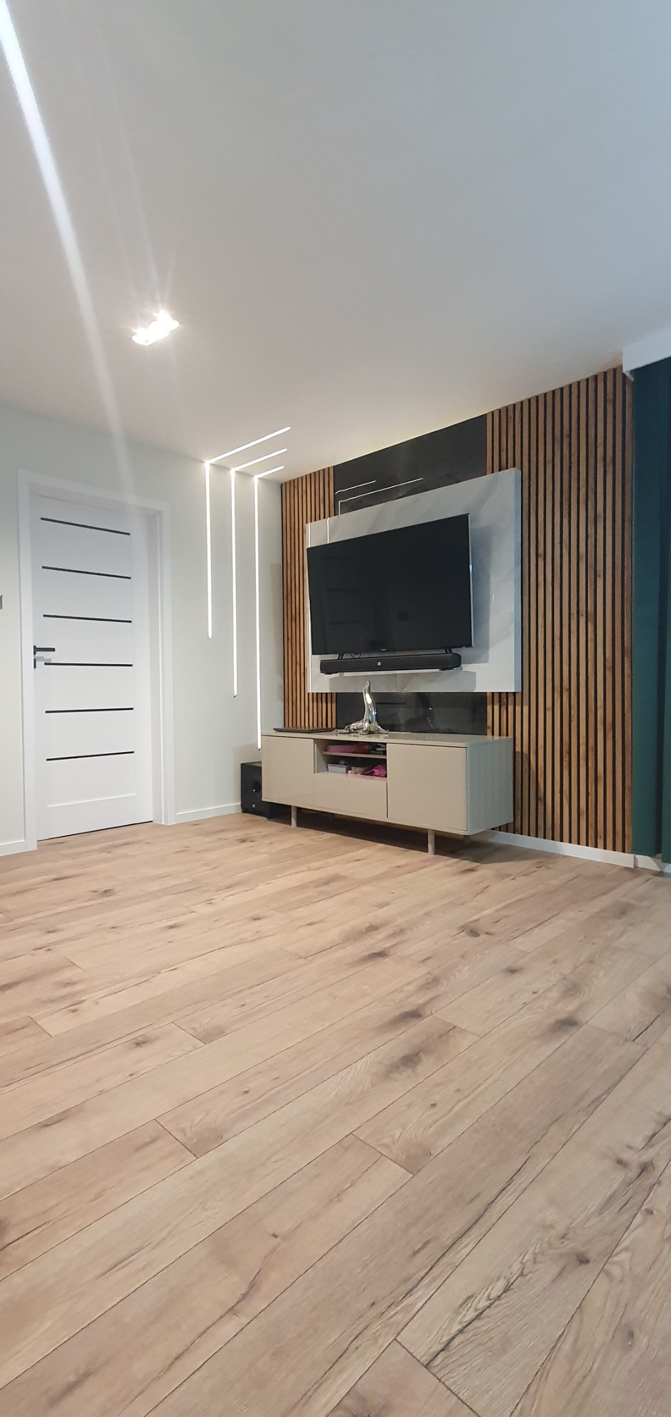 Salon z drewnianą podłogą, telewizorem na ścianie z drewnianymi panelami i oświetleniem LED, minimalistyczna aranżacja.