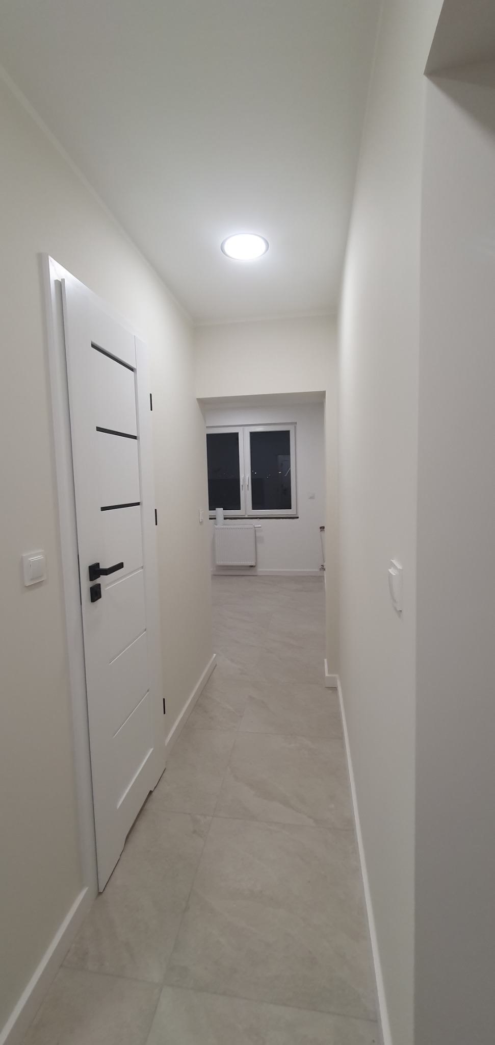 Jasny, wąski korytarz po remoncie z białymi ścianami, płytkami podłogowymi w odcieniach szarości i beżu, białą, nowoczesną ościeżnicą z czarną klamką i widokiem na okno z grzejnikiem.