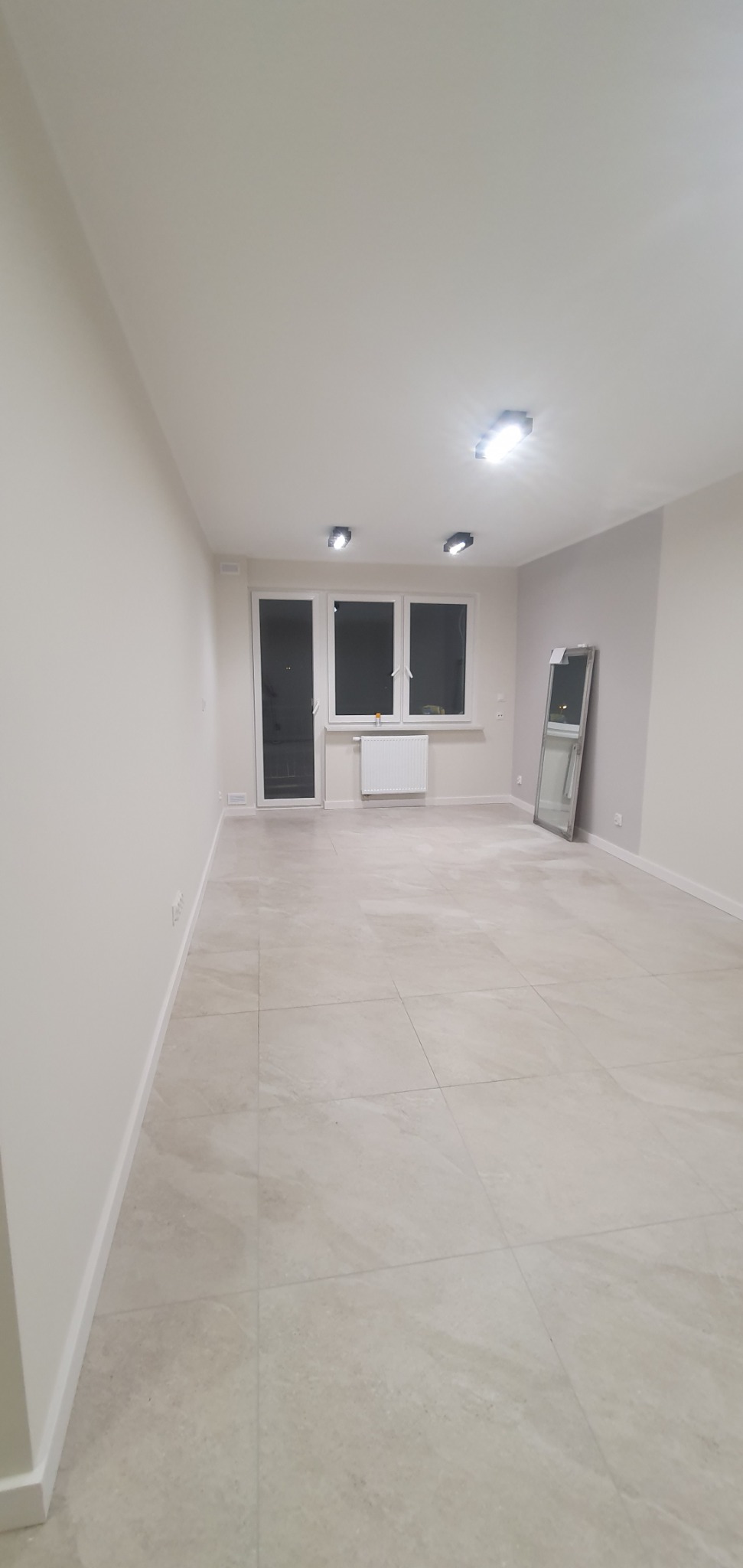 Puste, wykończone wnętrze pokoju z beżowymi płytkami podłogowymi, białymi ścianami, grzejnikiem pod oknem i lustrem opartym o ścianę.