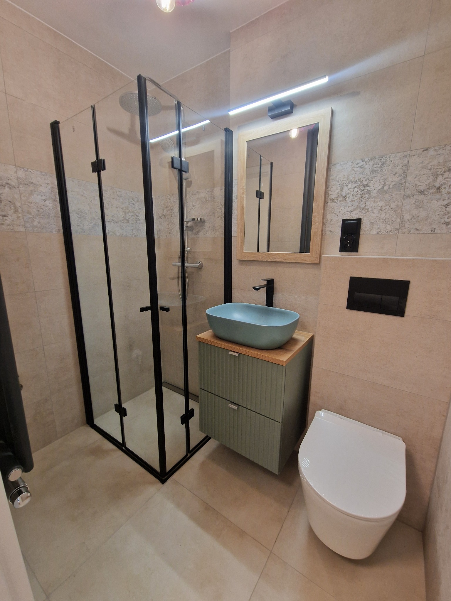 Nowoczesna łazienka z kabiną prysznicową typu walk-in z czarnymi profilami, umywalką nablatową w kolorze turkusowym na szafce z frezowanymi frontami, wiszącą toaletą i lustrem z oświetleniem LED.