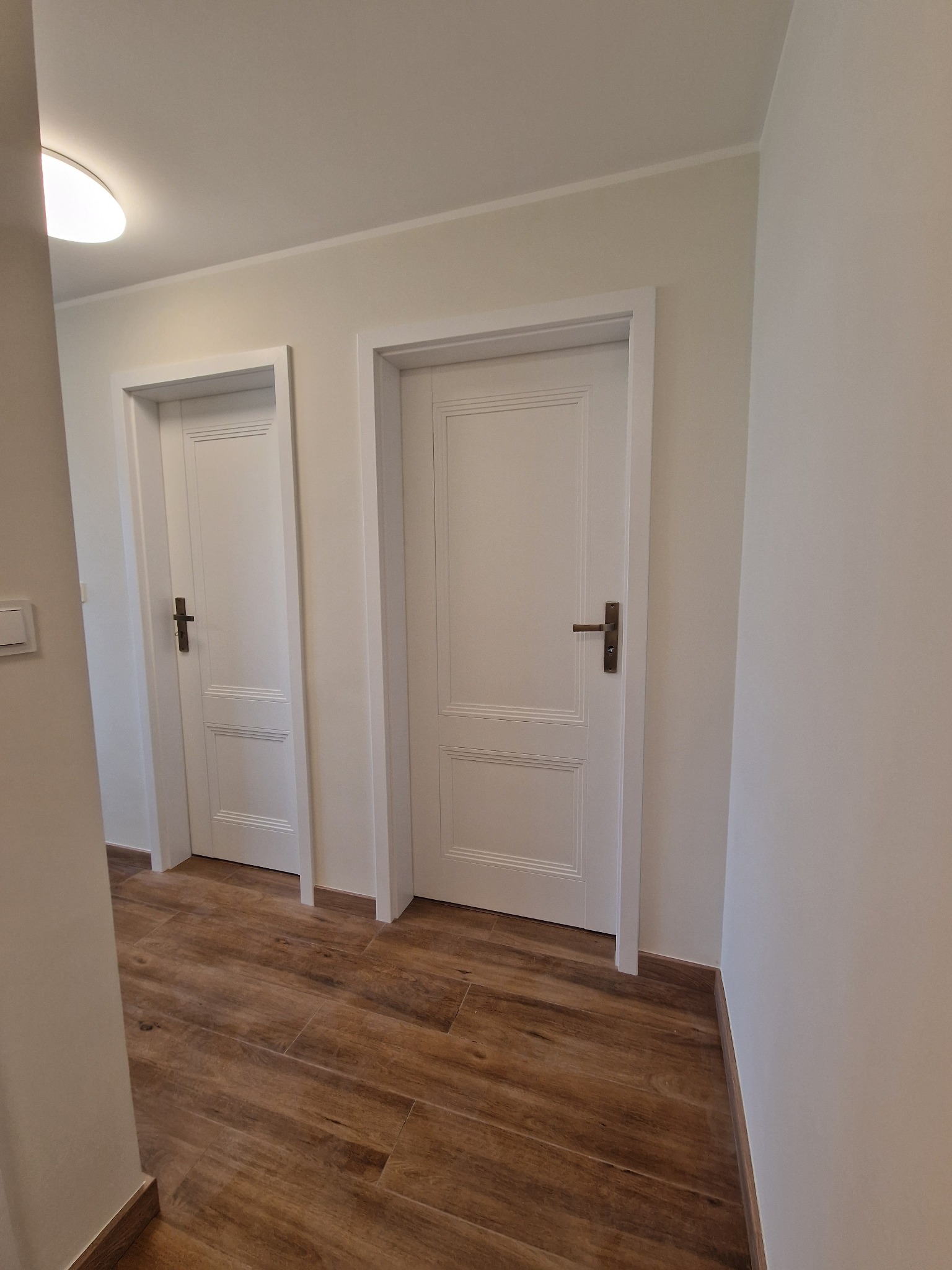 Korytarz z dwoma białymi drzwiami z ozdobnymi panelami i brązowymi klamkami, wykończony podłogą z płytek imitujących drewno.