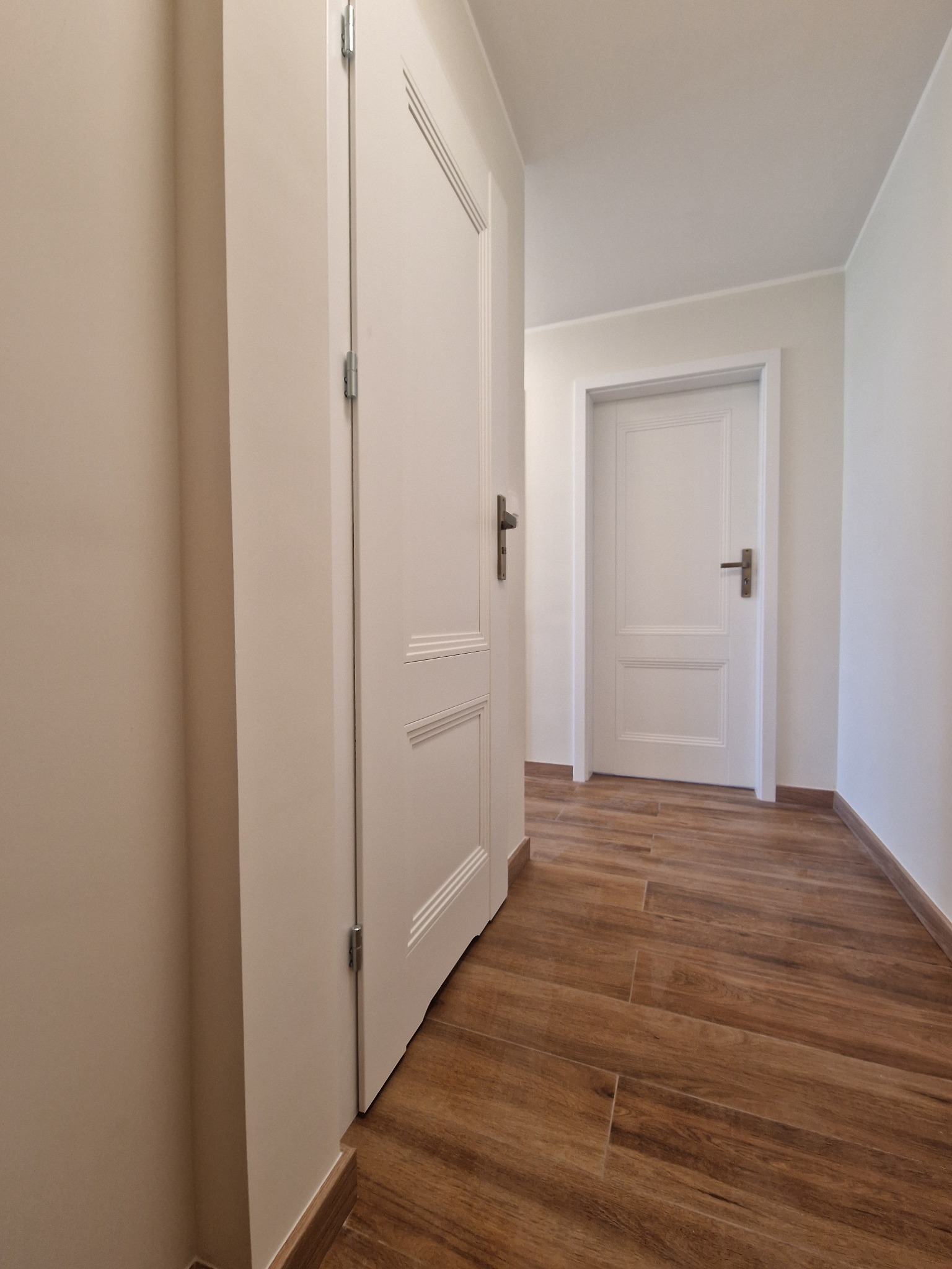 Korytarz z drewnopodobną podłogą, białymi ścianami i dwiema białymi, klasycznymi drzwiami z brązowymi klamkami; widok z niskiego kąta.