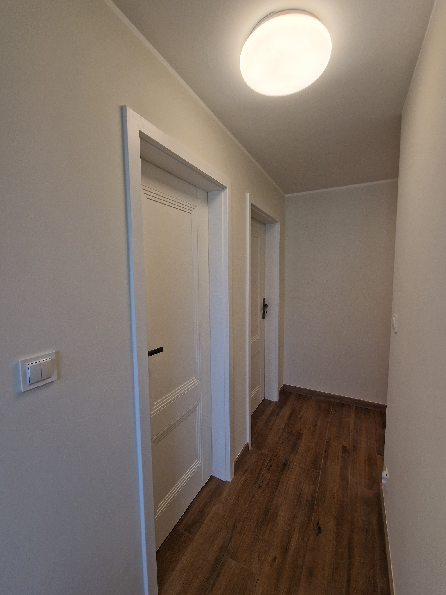 Korytarz po odnowieniu z dwiema parami białych drzwi, drewnopodobnymi panelami podłogowymi i okrągłą lampą sufitową.