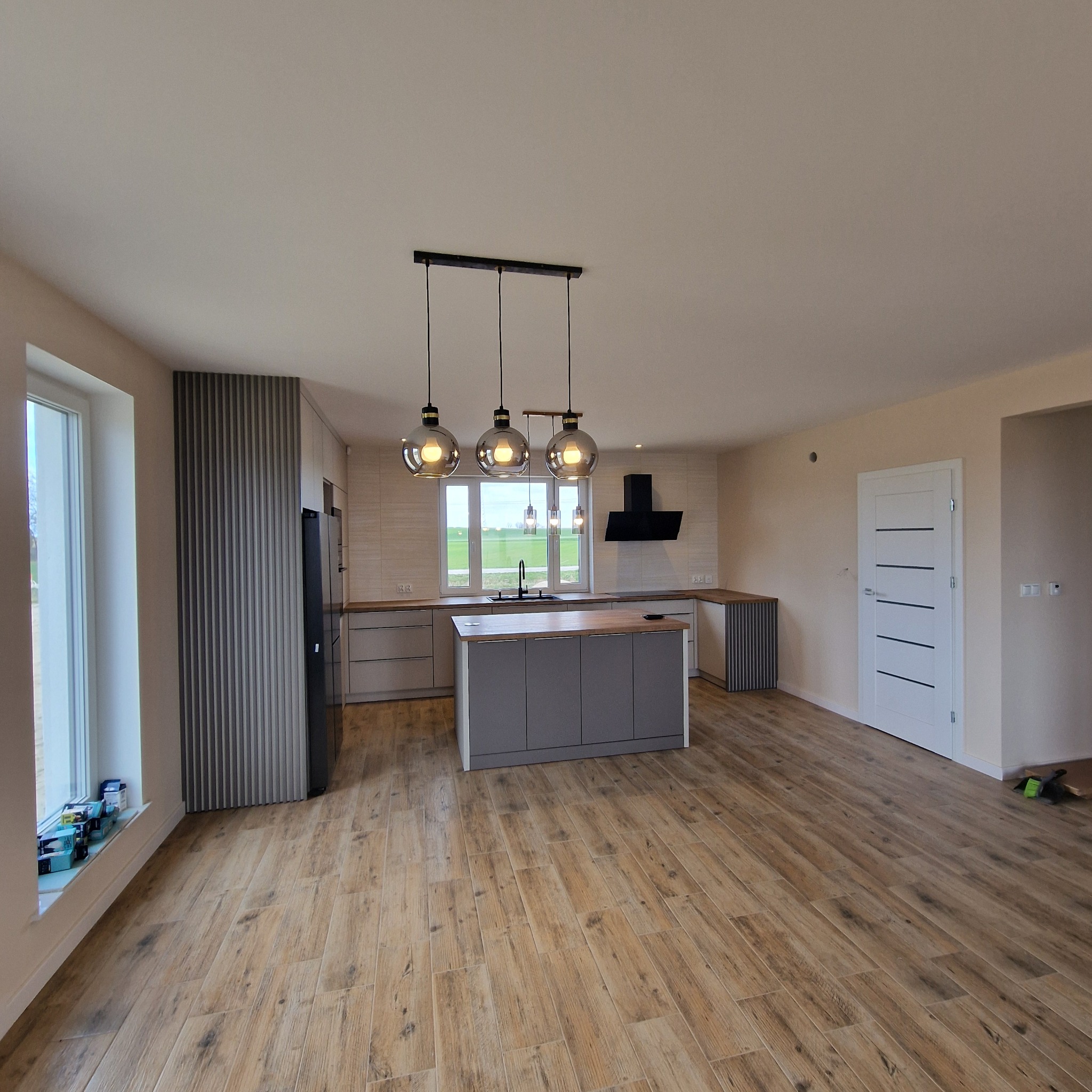Jasne wnętrze z drewnopodobną podłogą. Wyspa kuchenna z drewnianym blatem i wiszące, okrągłe lampy. W tle okno z widokiem na zieleń i białe drzwi.