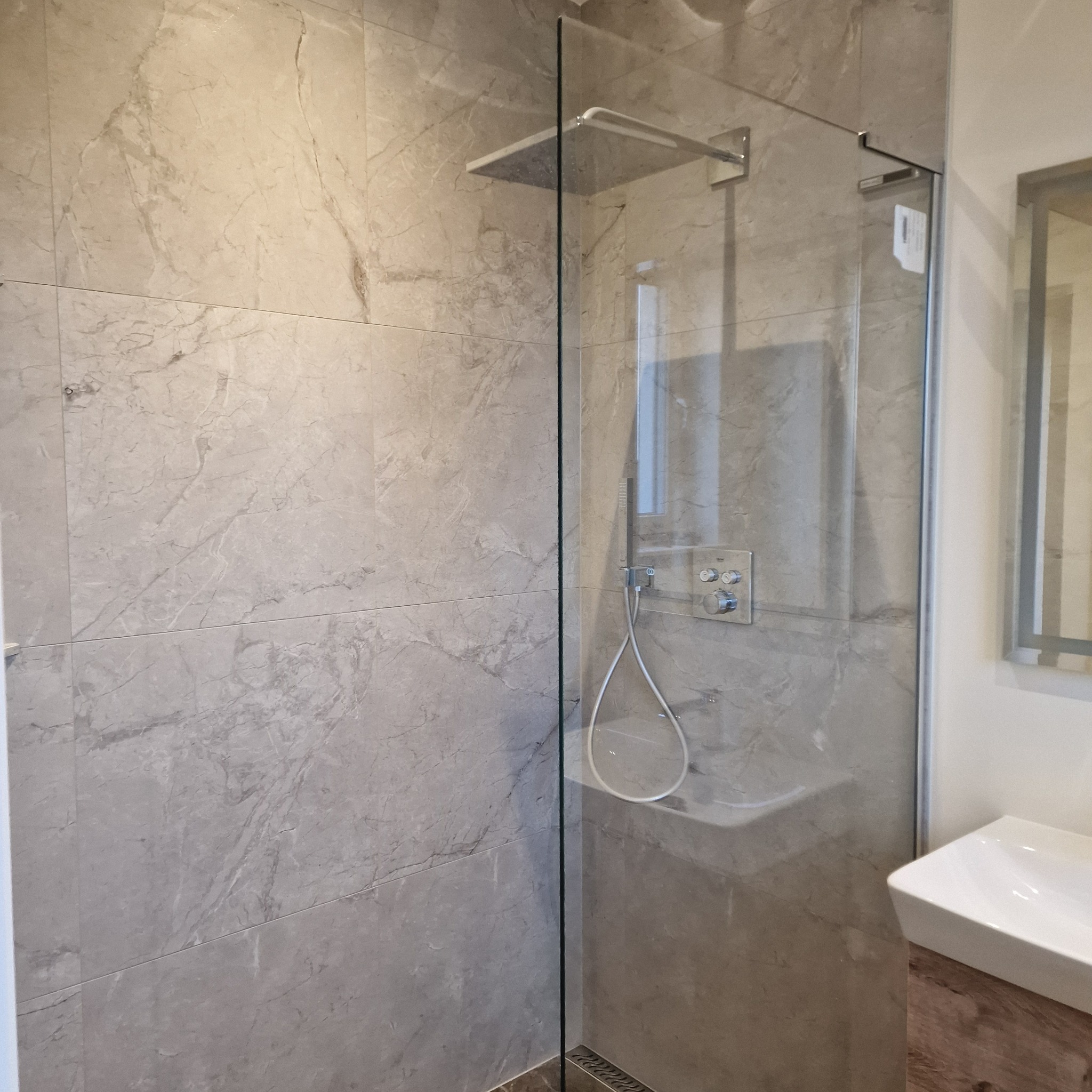 Nowoczesna kabina prysznicowa typu walk-in z szarymi płytkami imitującymi marmur, deszczownicą i minimalistyczną armaturą. Widoczny fragment umywalki.