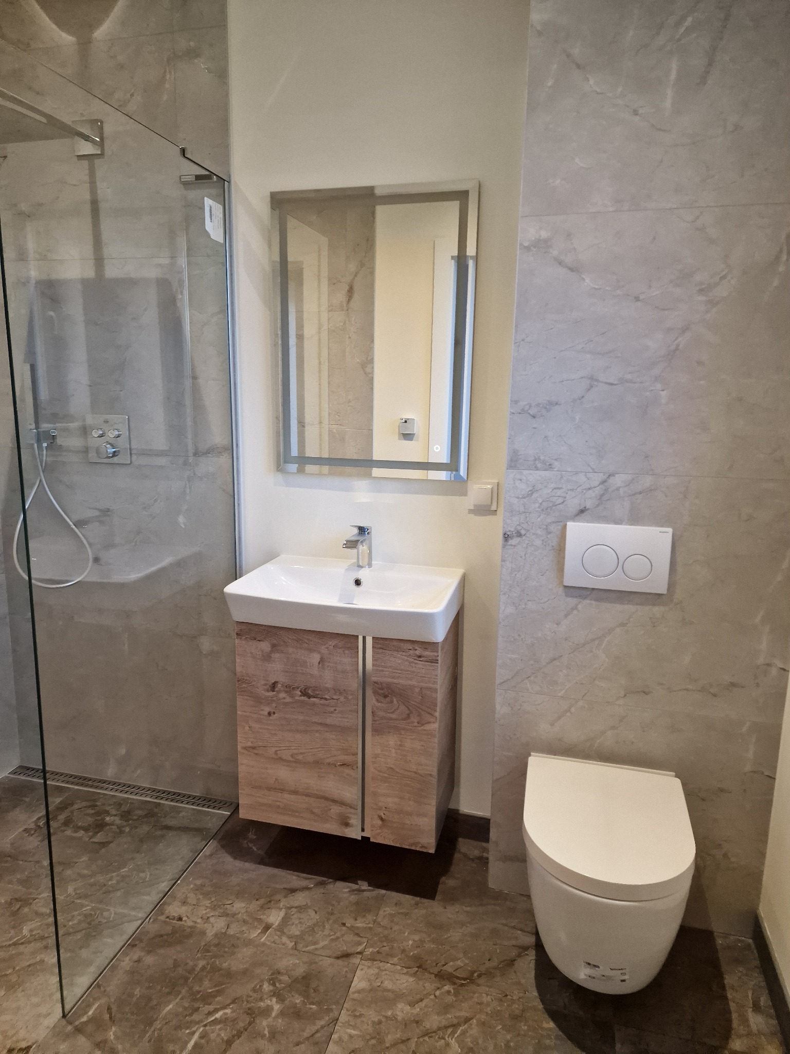 Nowoczesna łazienka z kabiną prysznicową, umywalką z szafką o drewnianym wzorze, podświetlanym lustrem i toaletą. Szare płytki imitujące marmur na ścianach i podłodze.