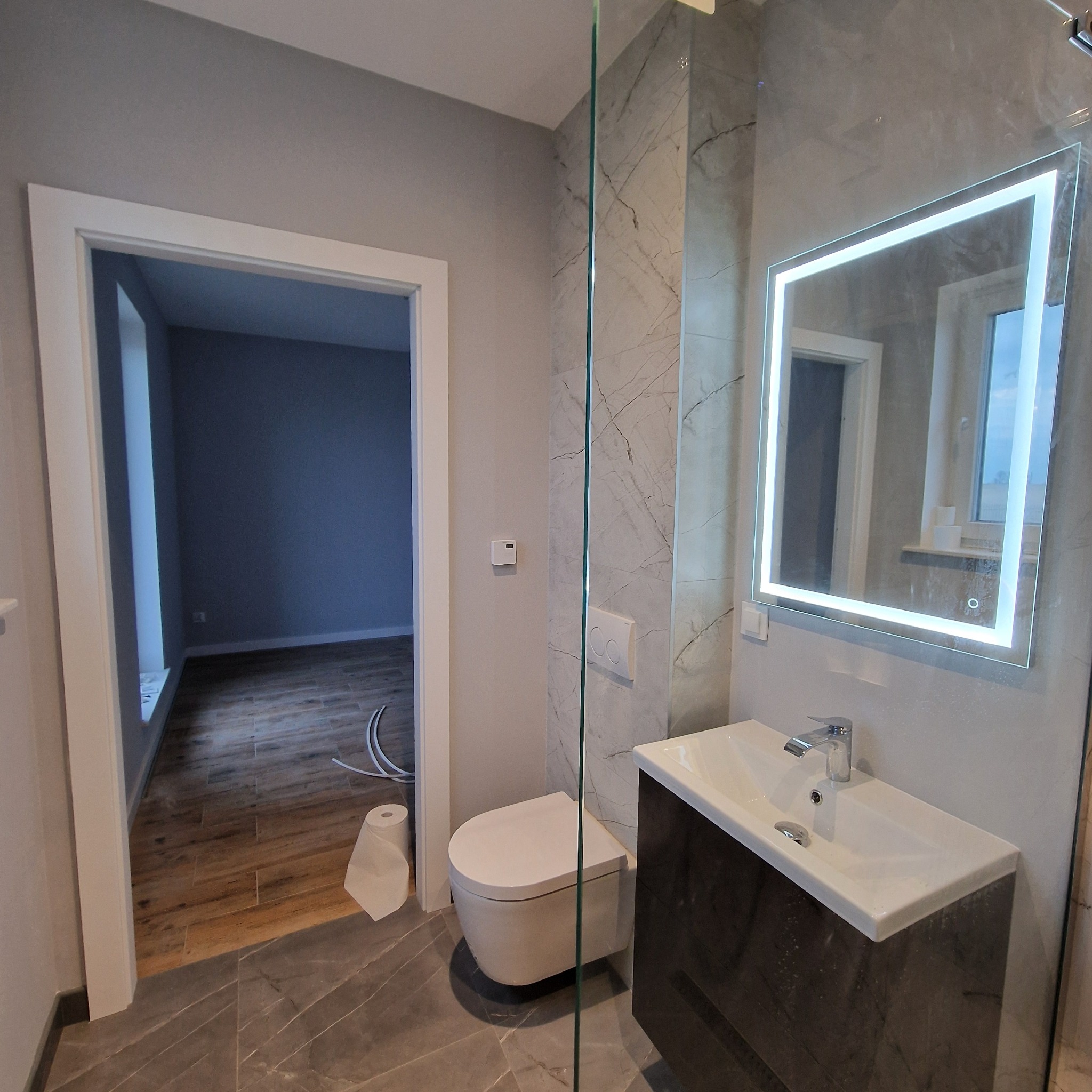 Nowoczesna łazienka po remoncie: podwieszana toaleta, umywalka z szafką i lustro z podświetleniem LED. Na podłodze i ścianach szare płytki imitujące marmur.