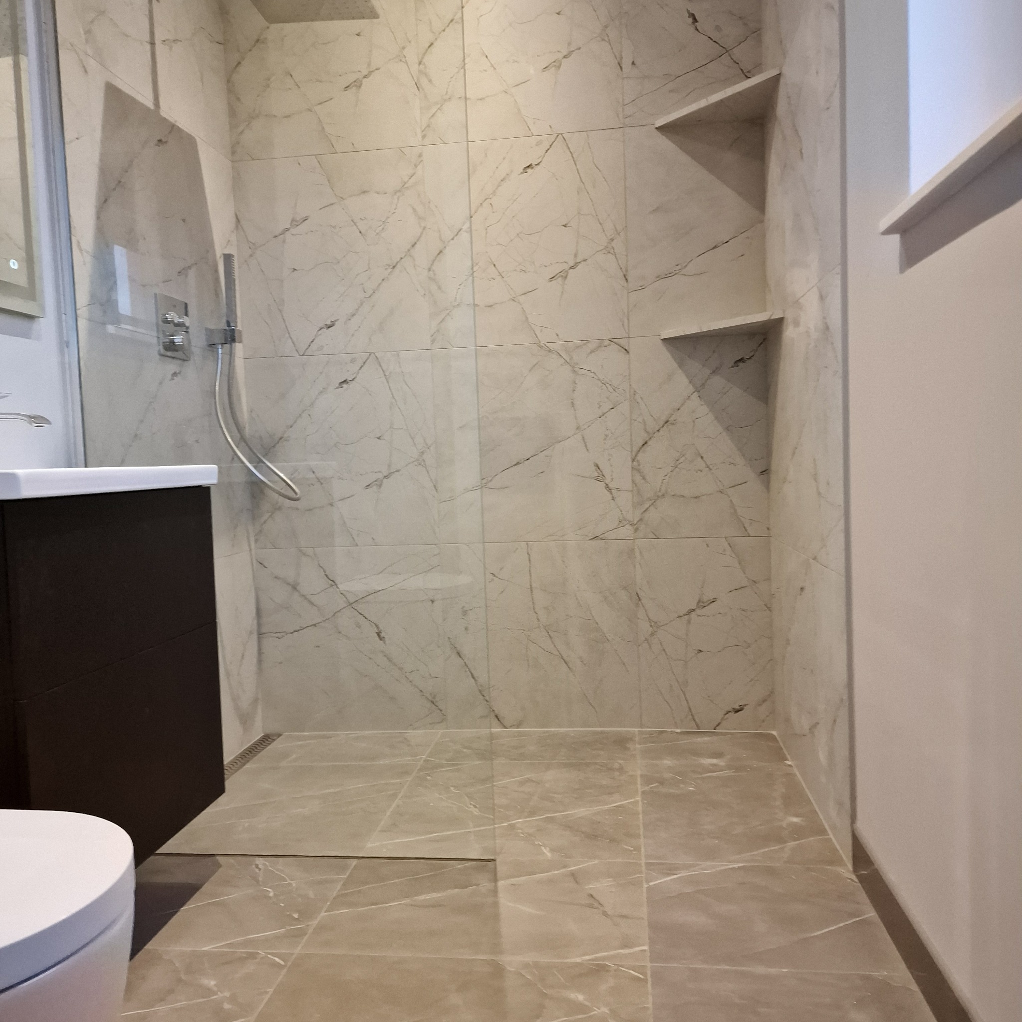 Wykończona łazienka z marmurowymi płytkami, kabiną prysznicową i wnękami na kosmetyki. Ciemna szafka pod umywalką i odpływ liniowy.