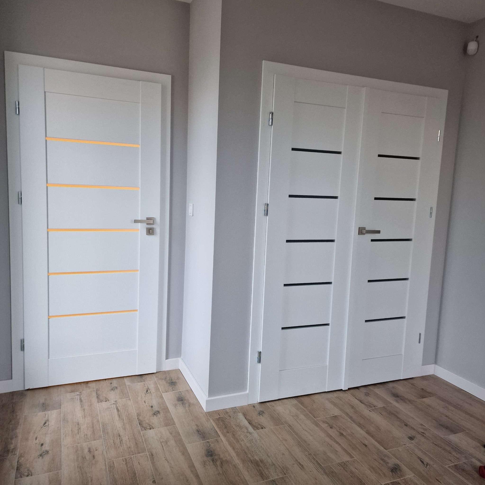 Dwa rodzaje białych drzwi wewnętrznych z poziomymi frezami (żółte i czarne), zamontowane w szarej ścianie na tle podłogi z paneli imitujących drewno.