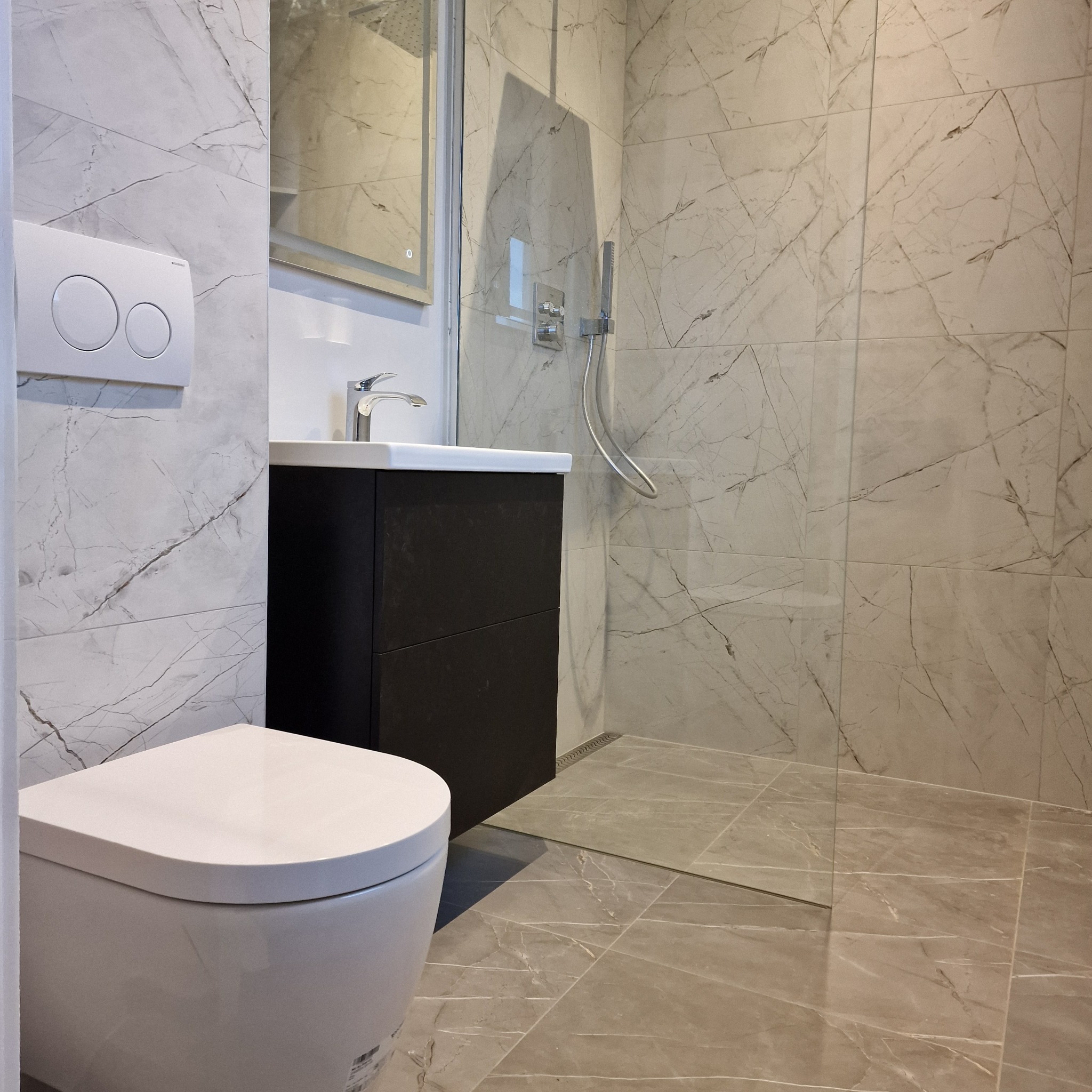 Nowoczesna łazienka z marmurowymi płytkami, wiszącą toaletą i kabiną prysznicową typu walk-in. Umywalka z szafką w kolorze ciemnego drewna. Minimalistyczny design.