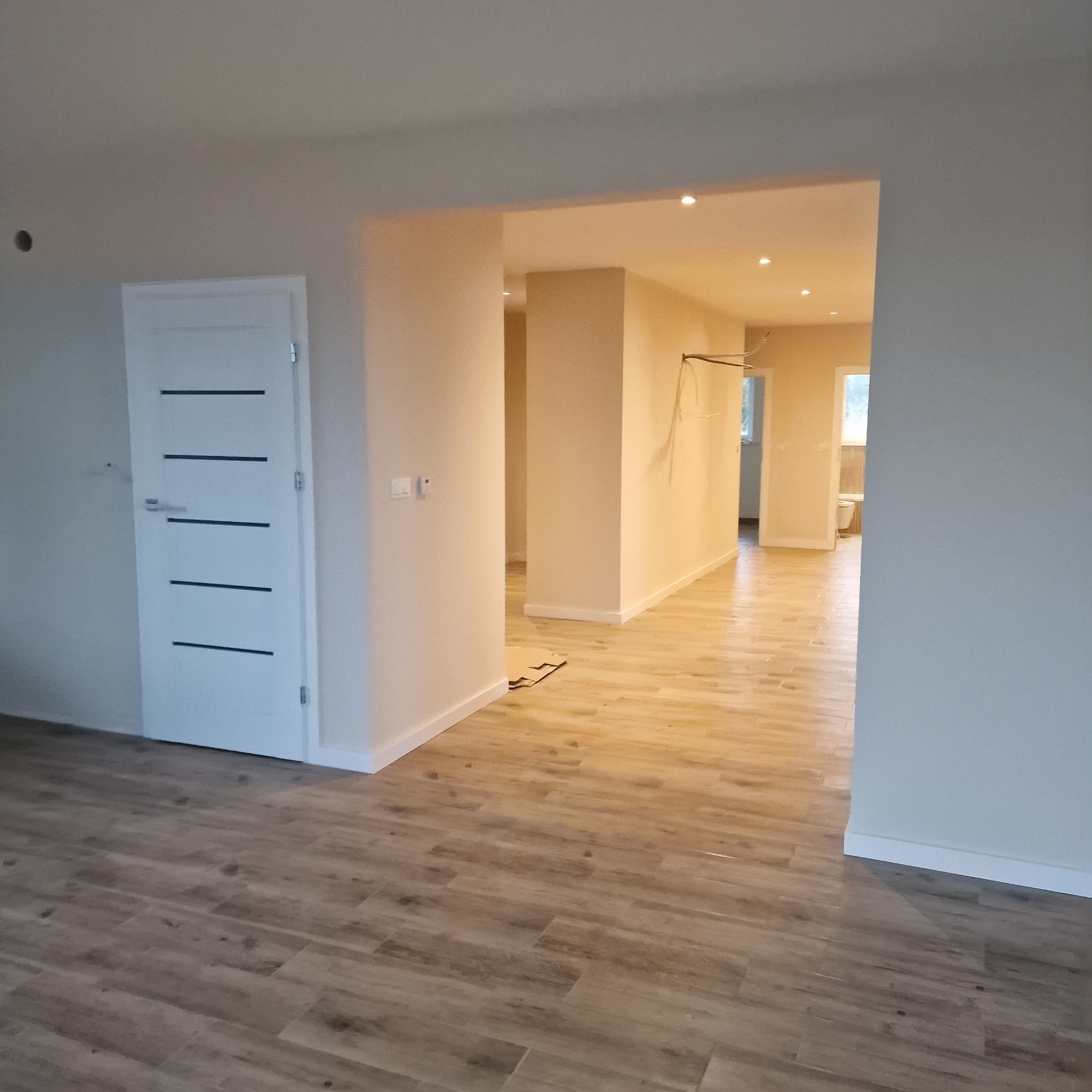 Jasne, przestronne wnętrze po remoncie z drewnopodobną podłogą i nowoczesnymi drzwiami. Widok na korytarz i łazienkę w głębi.