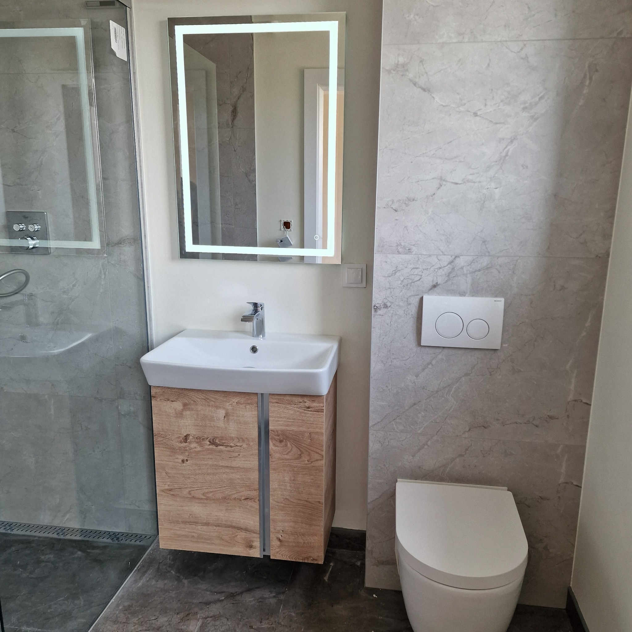 Nowoczesna łazienka po remoncie: marmurowe płytki, podświetlane lustro nad umywalką z drewnianą szafką, wisząca toaleta. Minimalistyczny design z szarą podłogą.