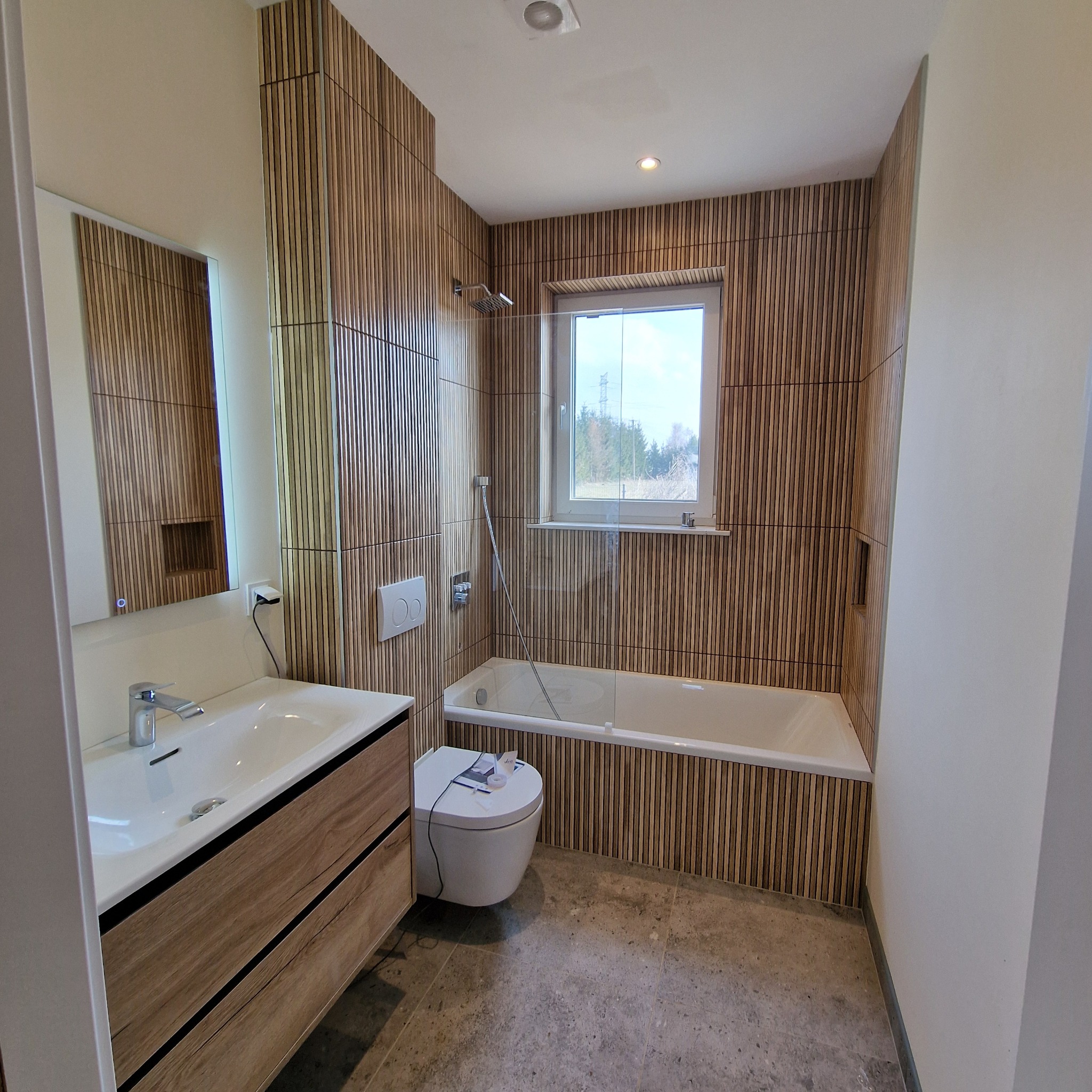 Nowoczesna łazienka z drewnianą okładziną ścian, białą armaturą i oświetlonym lustrem. Widok na okno z widokiem na zewnątrz. Umywalka z szafką z drewnopodobnym fornirem.