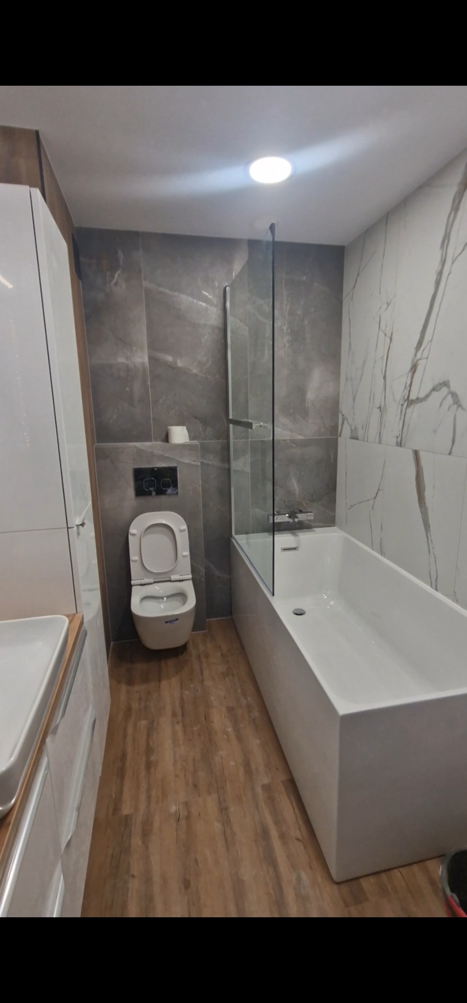 Nowoczesna łazienka po remoncie: biała wanna z parawanem, podwieszana toaleta i drewnopodobne panele podłogowe. Minimalistyczny design z szarymi i białymi płytkami.