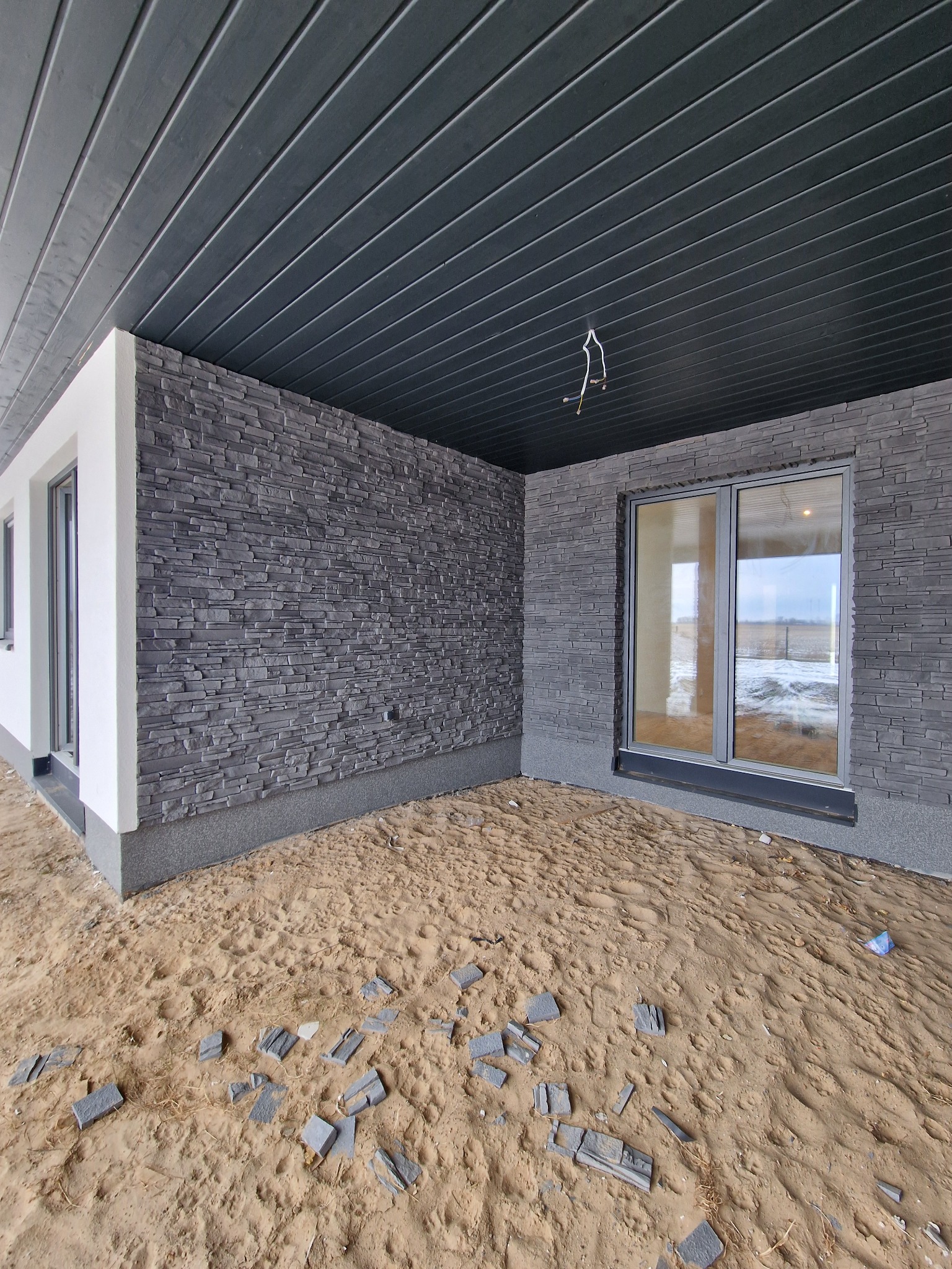 Montaż elewacji z kamienia dekoracyjnego na budynku z szarą podbitką. Na ziemi leżą fragmenty płytek. Widoczne okno i instalacja elektryczna.