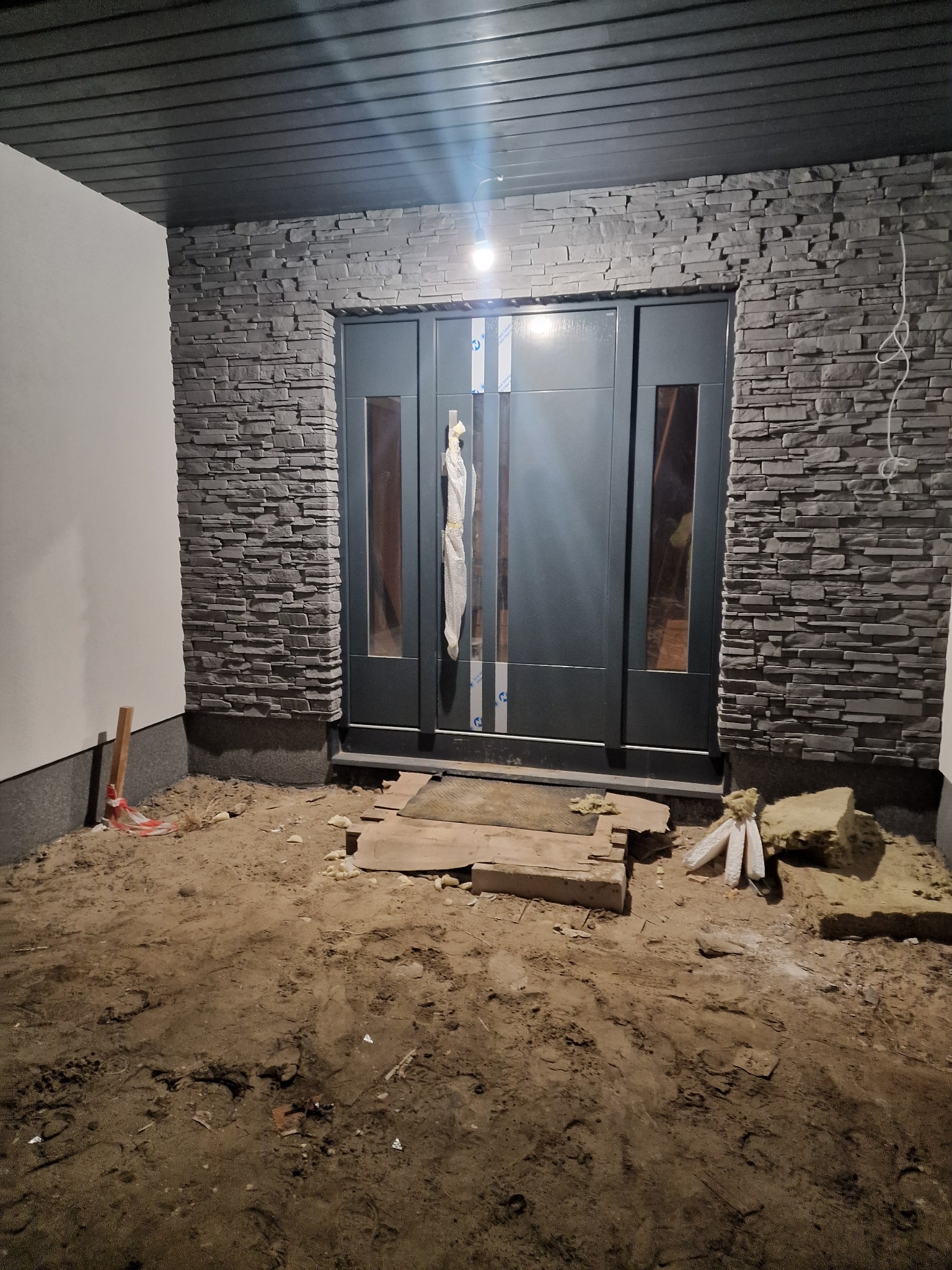 Montaż szarych drzwi wejściowych z przeszkleniami, obłożonych kamieniem dekoracyjnym na elewacji domu. Widoczne materiały budowlane i ziemia.