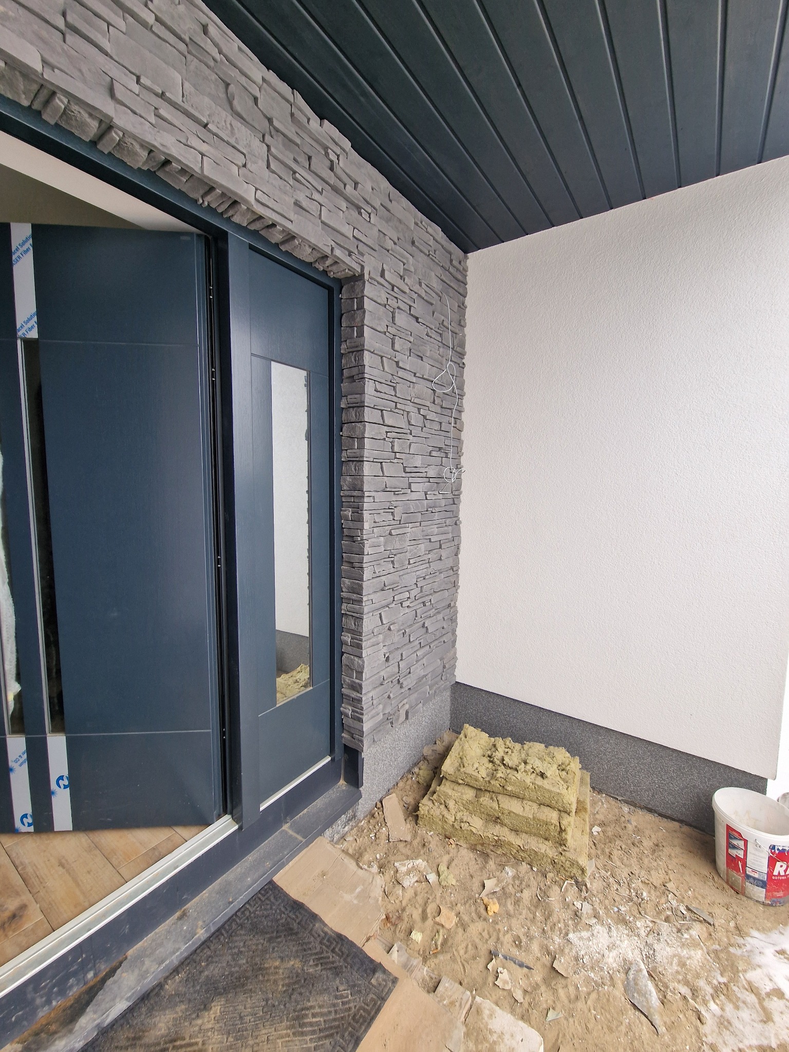 Montaż drzwi zewnętrznych z kamienną okładziną elewacyjną. Widoczny etap prac, izolacja i niebieskie drzwi z szybą. Wykończenie w nowoczesnym stylu.