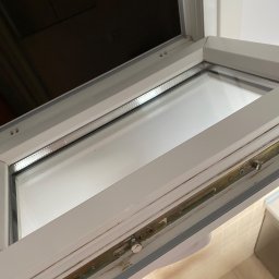 Pszczółka Cleaning - Otwarta, brudna rama okienna PCV z widocznymi śladami po czyszczeniu i metalowymi okuciami.