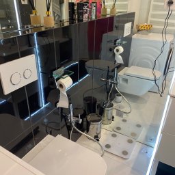 Pszczółka Cleaning - Nowoczesna łazienka z czarnymi płytkami i białymi elementami wyposażenia, widoczne toalety podwieszane, pralka, waga łazienkowa oraz aromatyczne dyfuzory zapachowe na półce.