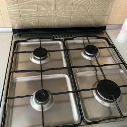 Czteropalnikowa kuchenka gazowa ze stali nierdzewnej, widok z góry, czyste palniki i ruszt, beżowe płytki na ścianie w tle.