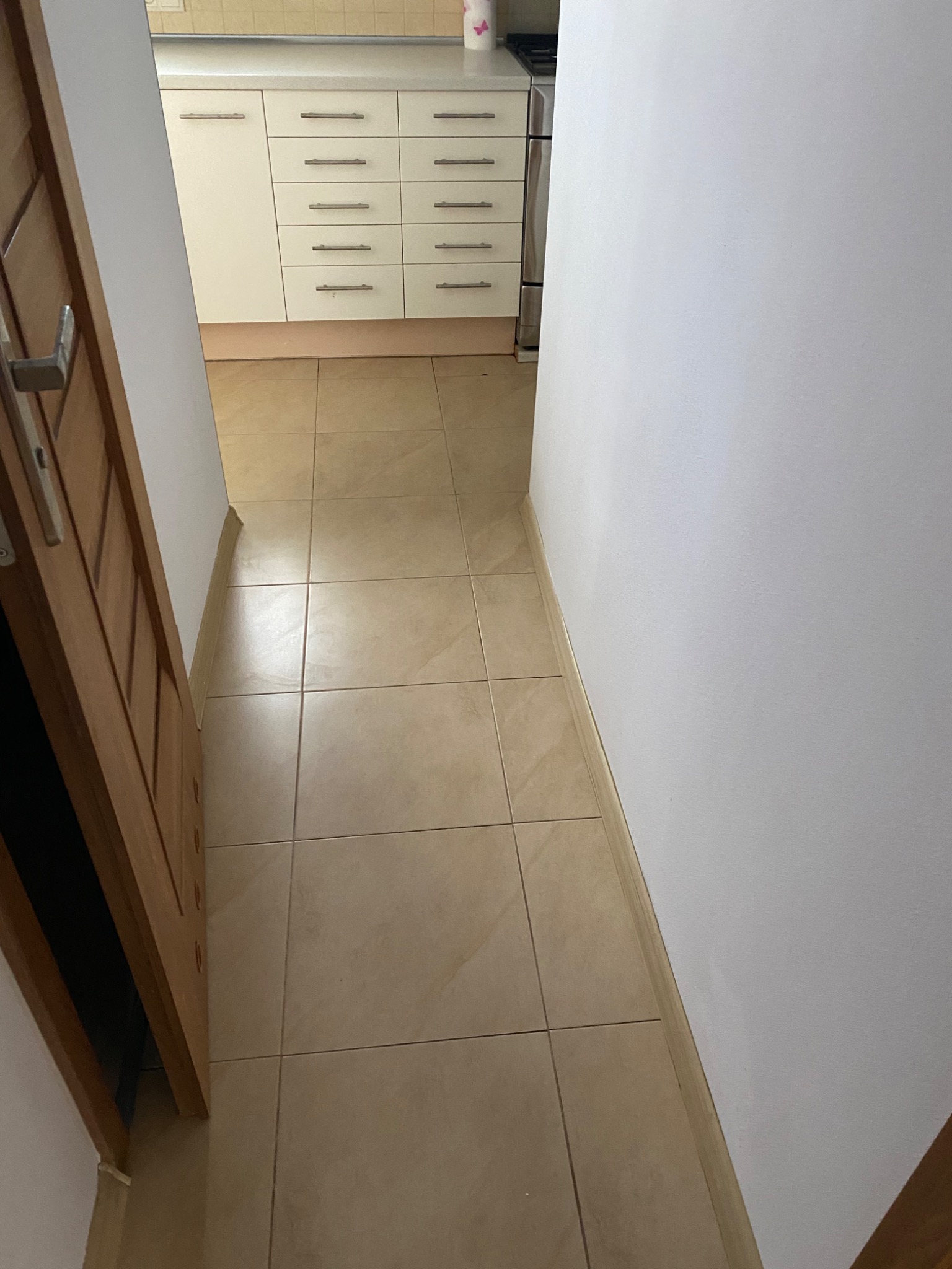 Wnętrze mieszkania z beżowymi płytkami na podłodze, widok na białe szafki kuchenne z metalowymi uchwytami przez otwarte drewniane drzwi.