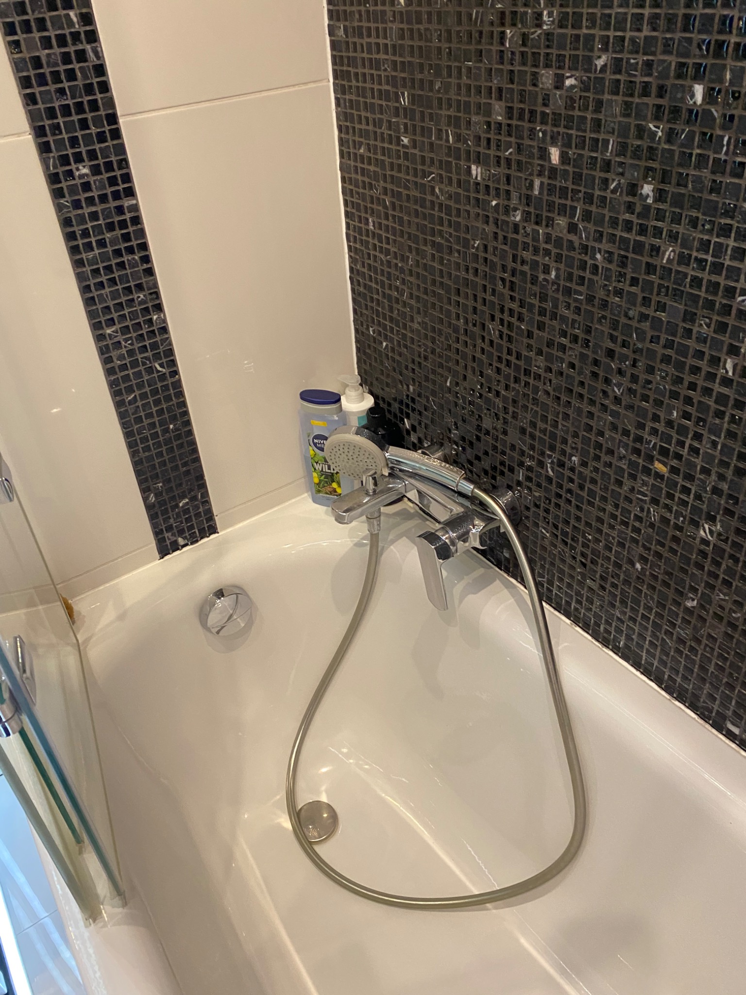 Biała wanna z chromowaną baterią i słuchawką prysznicową, obok butelki kosmetyków, na tle ściany wyłożonej czarno-białą mozaiką.