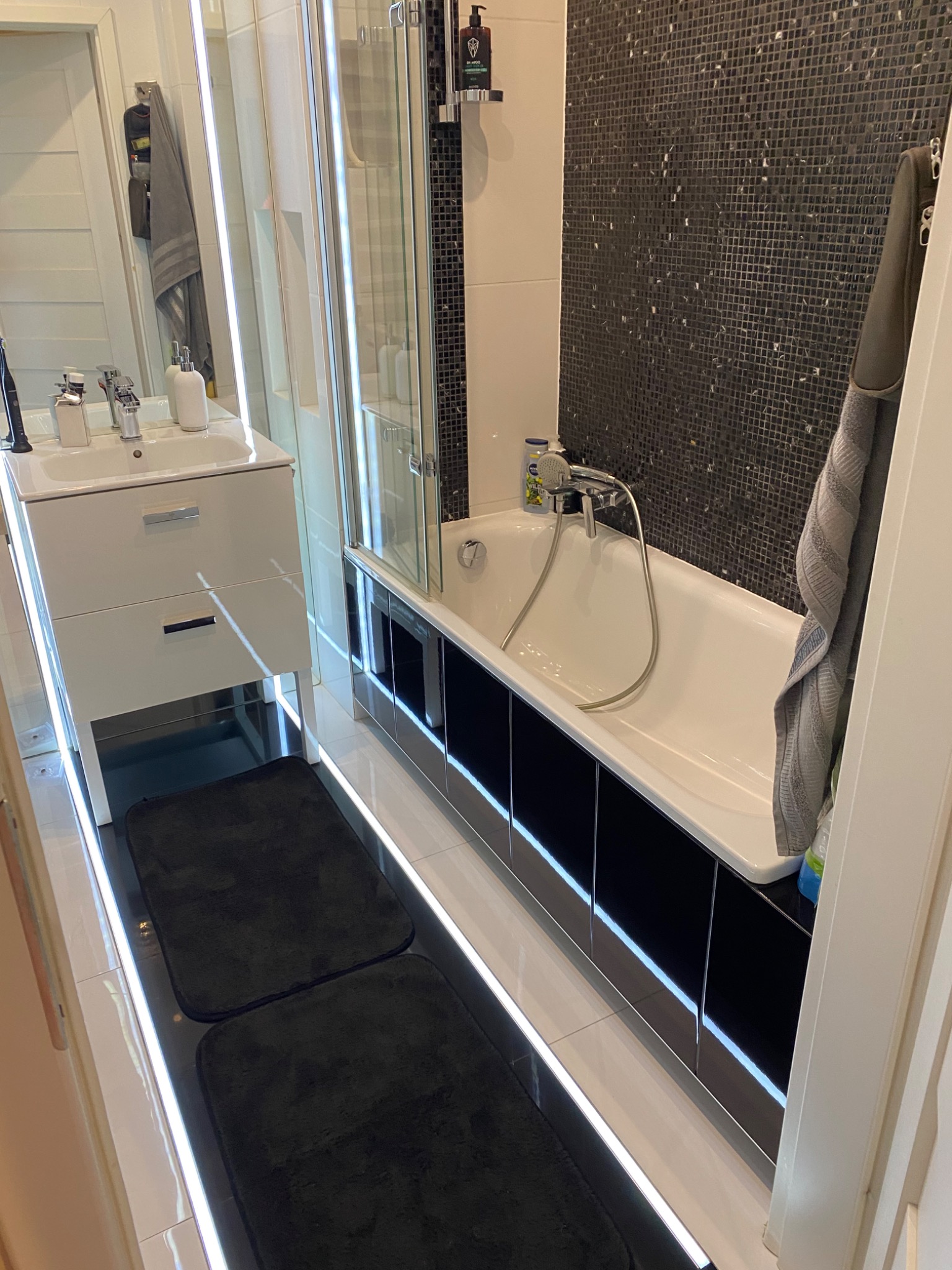 Nowoczesna łazienka z białą szafką pod umywalką, wanną z czarnymi panelami i mozaiką na ścianie, oświetlona listwami LED