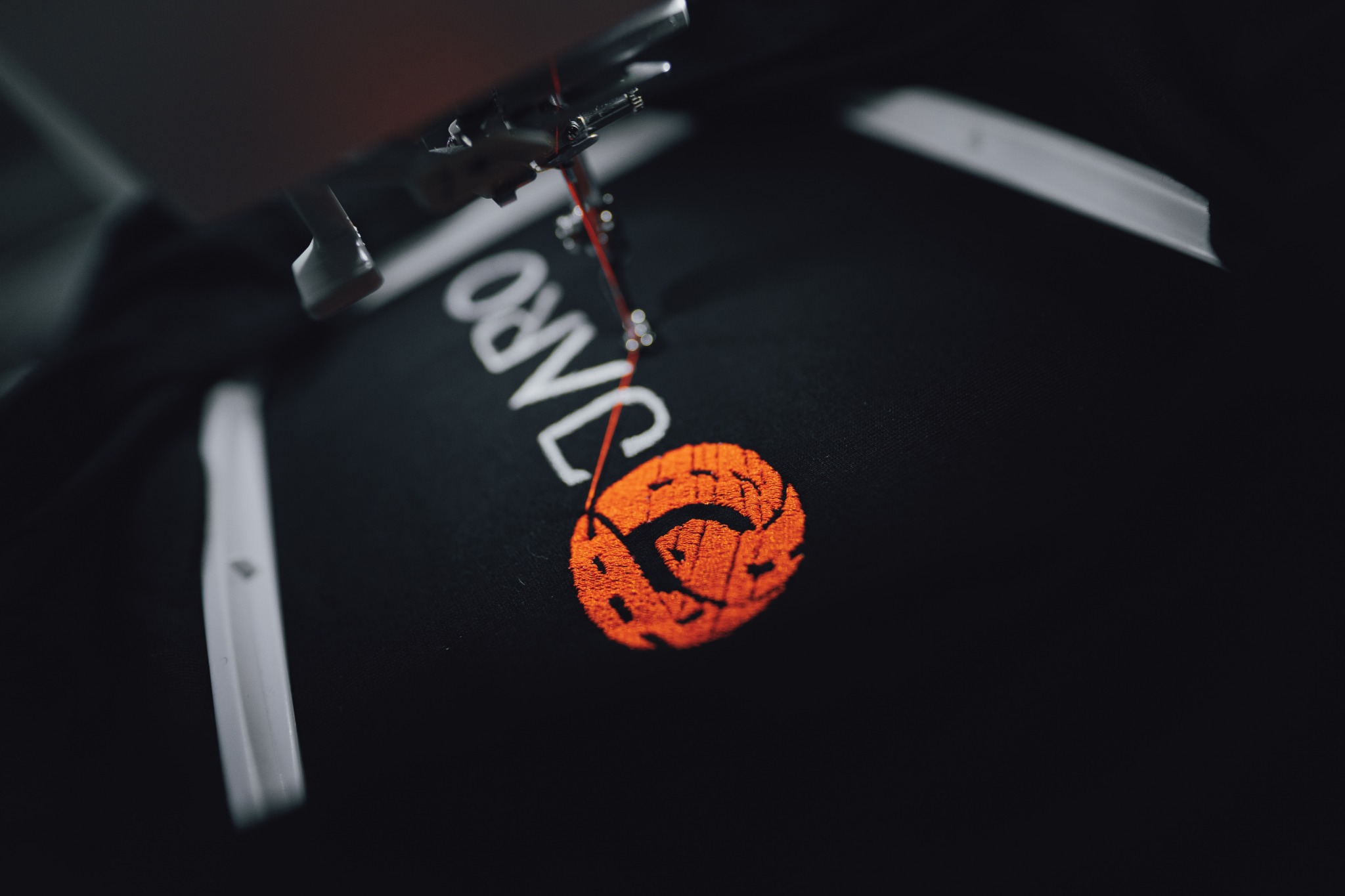 Proces haftowania pomarańczowego logo na czarnym materiale, widoczna igła maszyny i nitka.