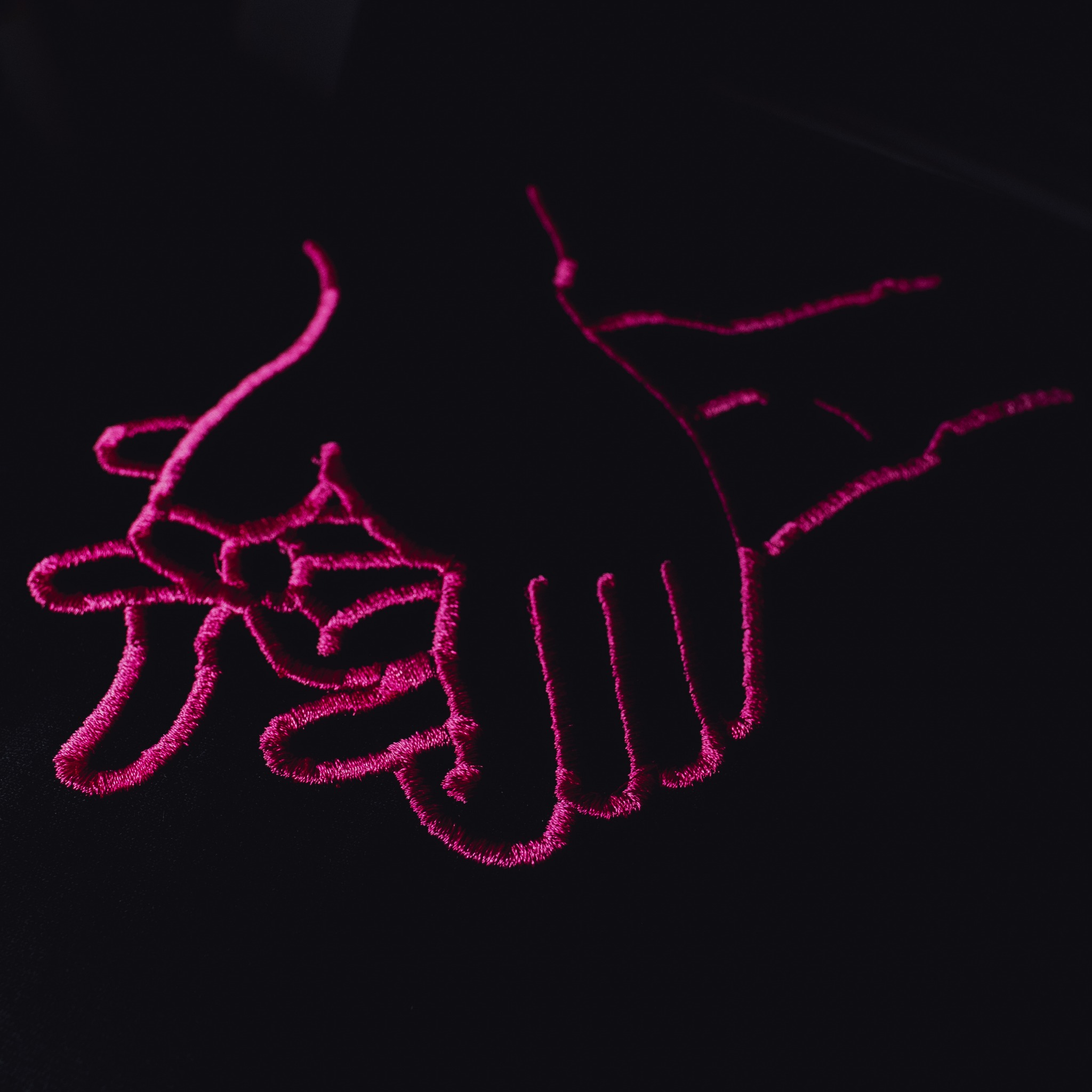 Zbliżenie na czarną tkaninę z wyhaftowanym różowym konturem dwóch dłoni, tworzących symboliczny uścisk.