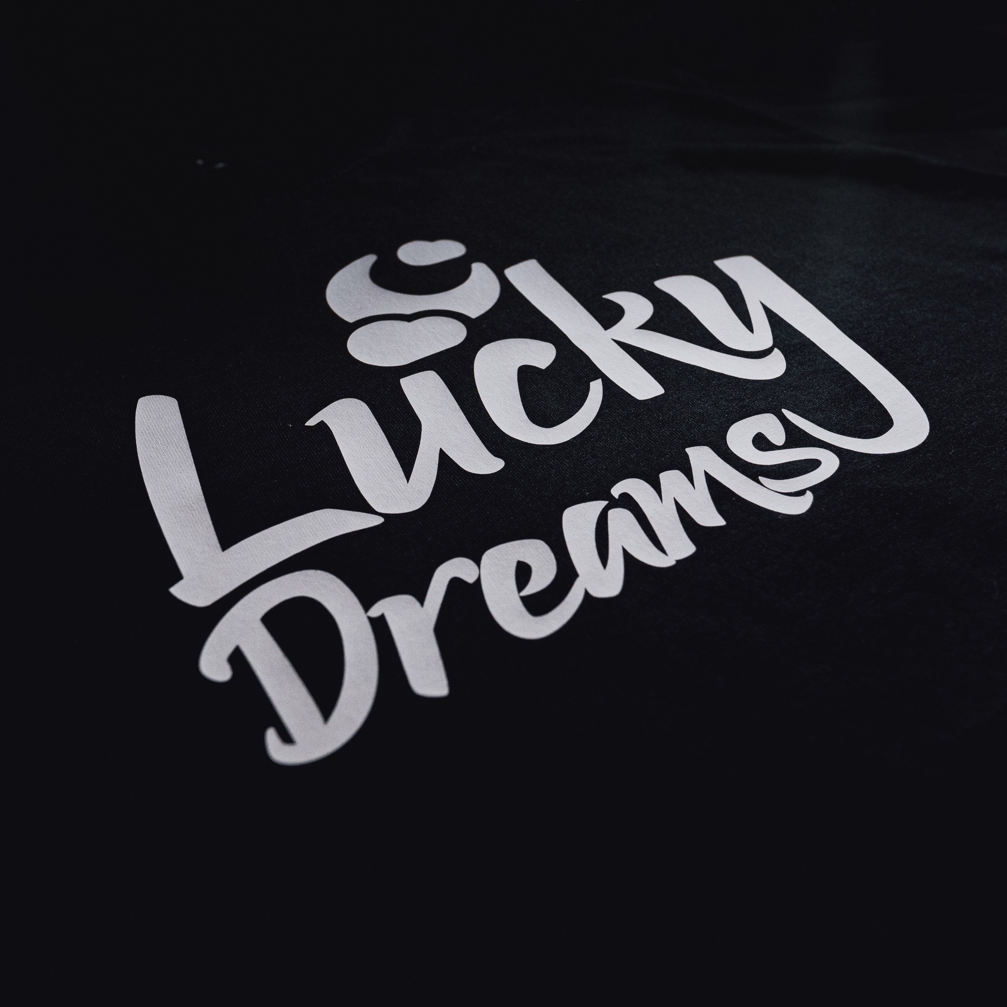 Czarna tkanina z białym, zakrzywionym napisem 'Lucky Dreams' pod kątem, oświetlona z góry.