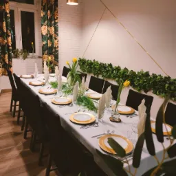 Uroczysty stół nakryty białym obrusem, z dekoracyjnymi złotymi talerzami, sztućcami, szklankami, serwetkami i wazonami z żółtymi tulipanami, oświetlony ciepłym światłem, z dekoracją ze sztucznych...