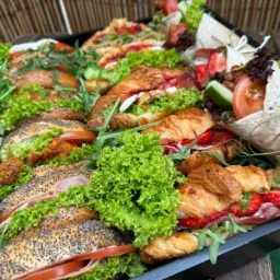 Taca z różnorodnymi kanapkami i wrapami, w tym bagietki z makiem, croissanty z szynką i truskawkami, oraz zawijasy z warzywami, przygotowana przez firmę z Radomia.