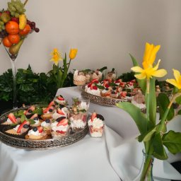 NALEŚNIKARNIA ANCYMONEK Małgorzata Wójcik - Stół z deserami: tartaletki z owocami, mini desery w kubeczkach, taca z ciastami, dekoracja z owoców w kielichu i żółte tulipany w wazonie.