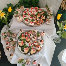 NALEŚNIKARNIA ANCYMONEK Małgorzata Wójcik - Elegancko zaaranżowany stół z deserami: mini bezy z owocami, tartaletki z kremem i owocami oraz desery w przezroczystych kubeczkach, udekorowane miętą, na srebrnych tacach, z kwiatami w tle.