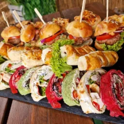Elegancko ułożone na czarnym talerzu mini burgery z wykałaczkami, roladki z kolorowych tortilli z różnymi nadzieniami oraz przekąski z ciasta francuskiego.