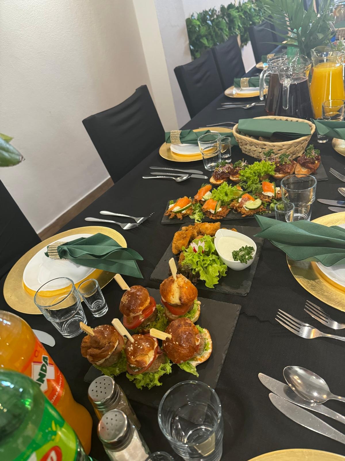 Elegancko zastawiony stół z czarnym obrusem, złotymi podkładkami, zielonymi serwetkami i różnorodnymi przekąskami: mini burgery na szpilkach, kanapeczki z łososiem, smażone kąski z sosem i sałatką,...