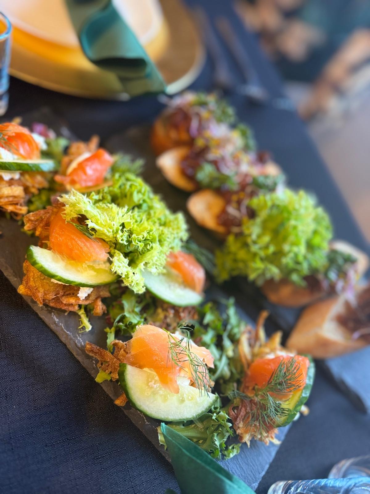 Elegancko podane przekąski na ciemnym talerzu: kanapeczki z łososiem, ogórkiem, sałatą i prażoną cebulką, oraz mini burgery z ciemnym sosem i zielenią, uzupełnione zieloną serwetką i kieliszkiem.