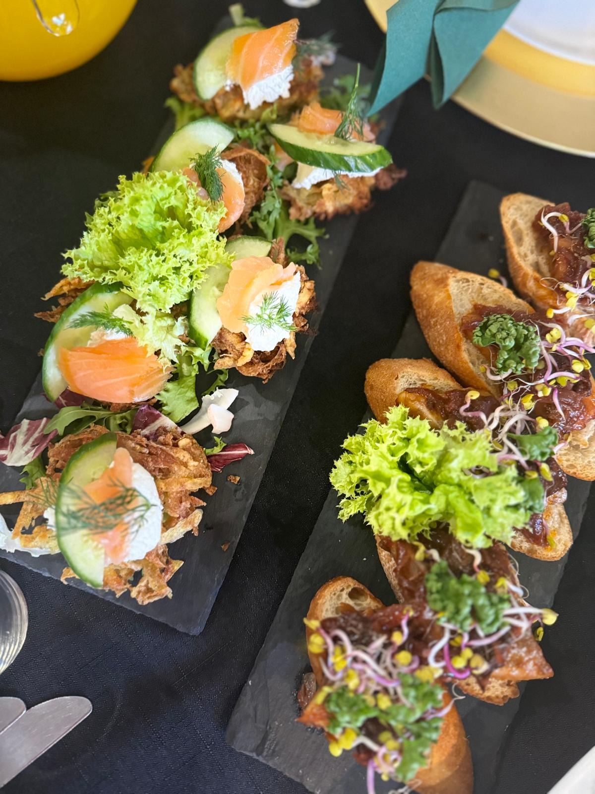 Dwie czarne deski z przekąskami: jedna z rosti z łososiem i ogórkiem, druga z bagietkami z konfiturą i kiełkami, udekorowane sałatą.