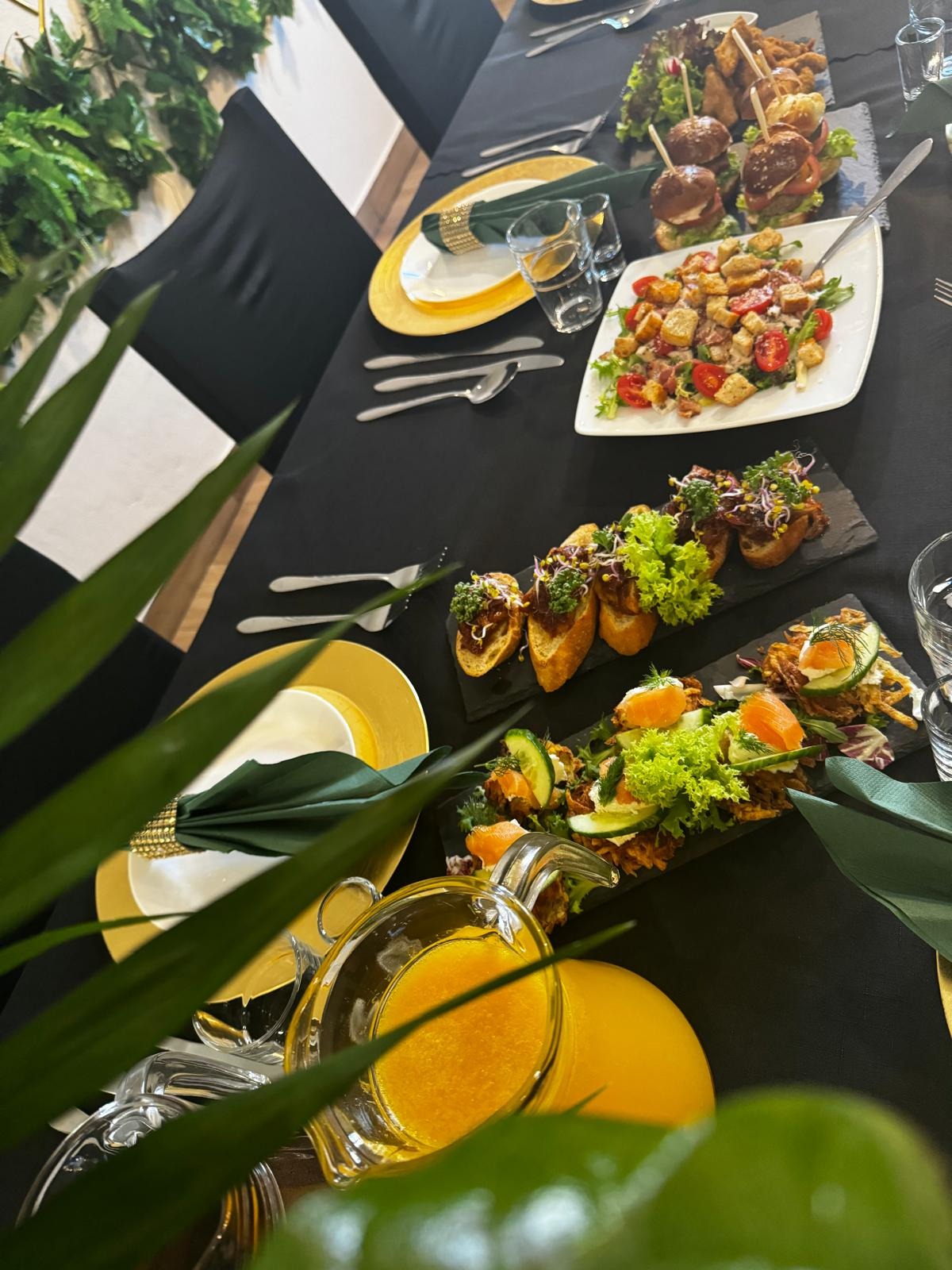 Elegancko zastawiony stół z czarnym obrusem, złotymi talerzami i zielonymi serwetkami, na którym znajdują się różnorodne przekąski: mini burgery na szpadkach, sałatka z grzankami, bruschetty...