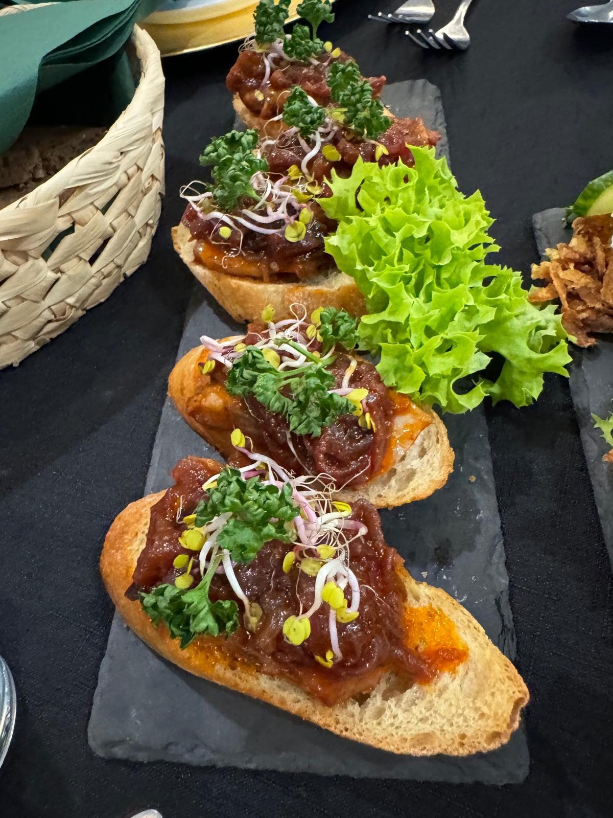 Bruschetta z ciemnym mięsem, kiełkami i zieleniną na ciemnej kamiennej płycie, obok koszyk z chlebem i sałata.